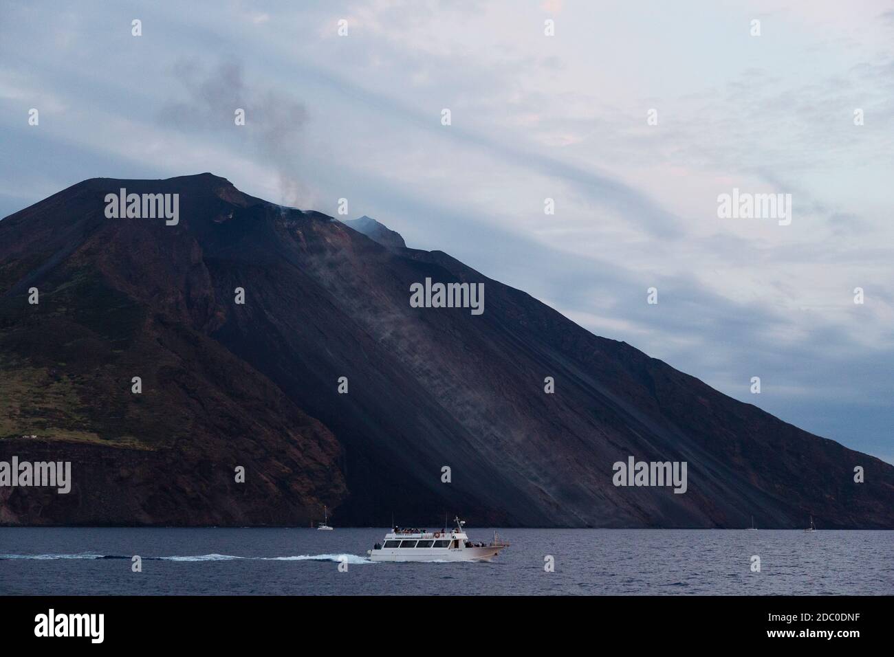 Sicilia, Italia. Una barca turistica passa di fronte all'isola vulcanica di Stromboli, mentre il vulcano attivo emette un scuro pennacchio di fumo e cenere. Foto Stock