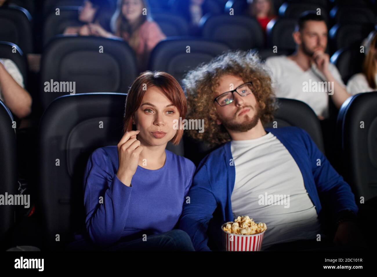 Fuoco selettivo della giovane coppia caucasica che guarda il film al cinema, seduto in comodi posti neri e che mangia il popcorn. Vista frontale di uomo in occhiali e donna con volti forti godendo di film. Foto Stock
