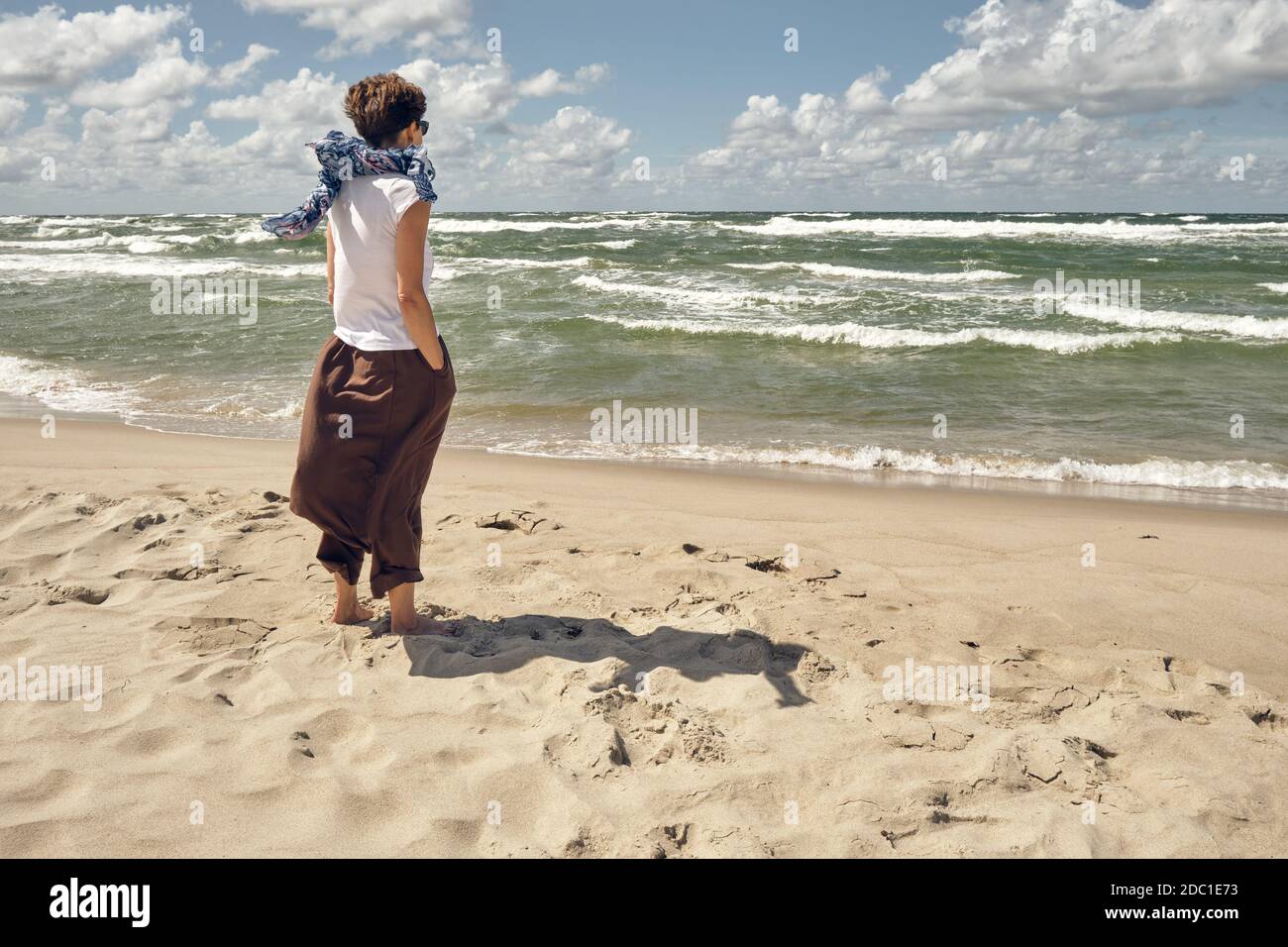 Donna di mezza età in piedi sulla spiaggia guardando ondeggiando mare in ventosa giornata estiva Foto Stock