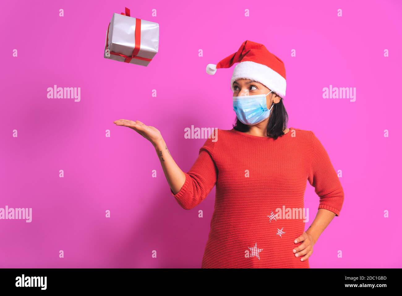 covid-19 concetto di natale. donna adulta felice con scatola regalo volante, maschera facciale, vestito babbo natale sorridente su sfondo rosa colorato. durino di natale Foto Stock