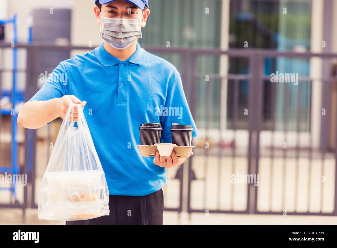 Giovane uomo asiatico di consegna in uniforme blu maschera di indossare faccia servizio di drogheria che dà riso scatole di plastica sacchetti di cibo e caffè in casa davanti sotto Foto Stock