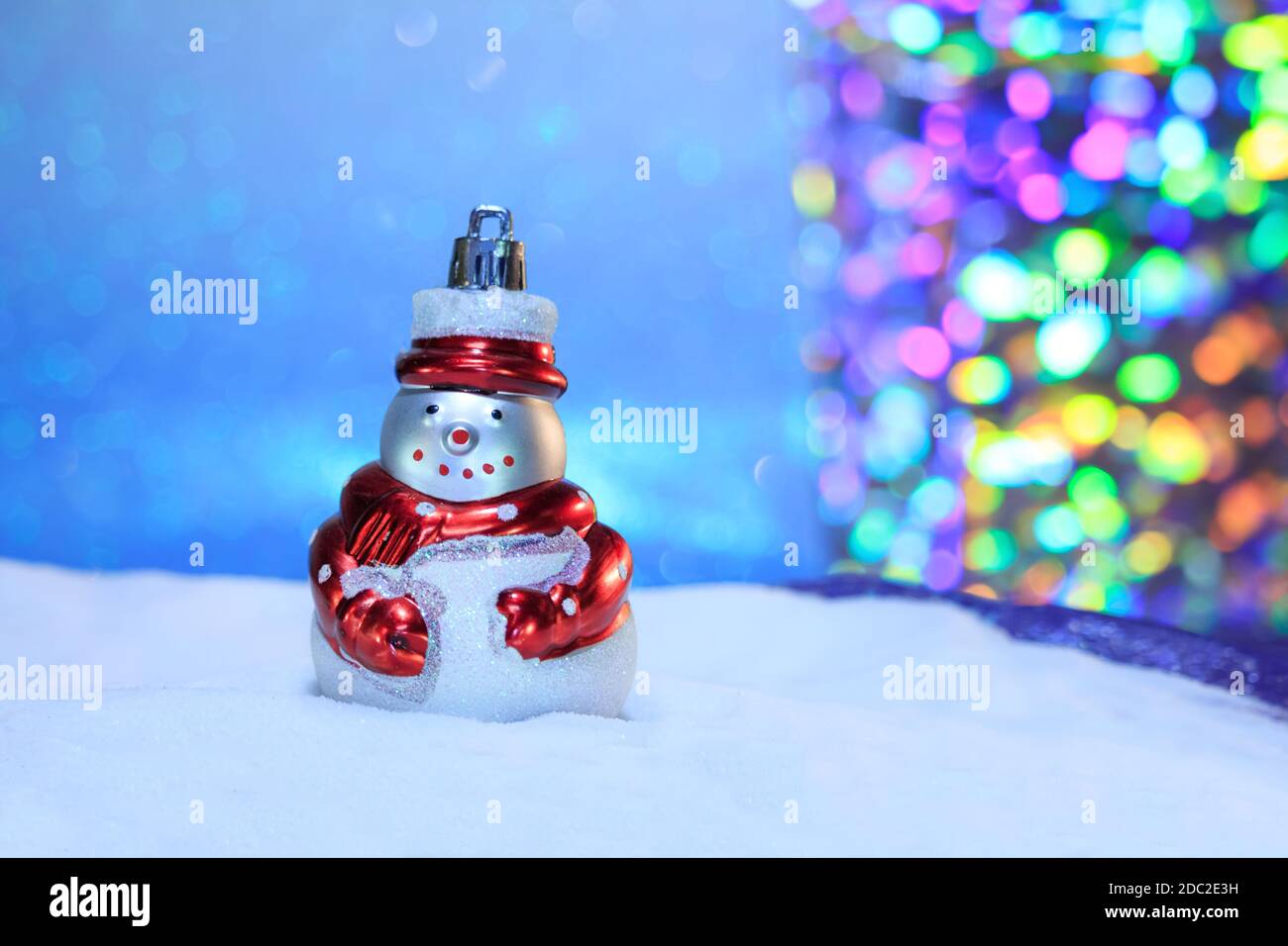 Pupazzo di neve con sciarpa rossa nella neve e cielo blu da dietro e bokeh multicolore. Sfondo sfocato. Buon Natale e carta di nuovo anno. Copia spa Foto Stock