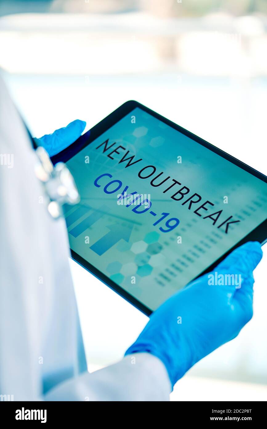 primo piano di un medico, indossando guanti chirurgici blu, avendo una tavoletta digitale in mano con il testo covid-19 nuovo focolaio nel suo schermo Foto Stock
