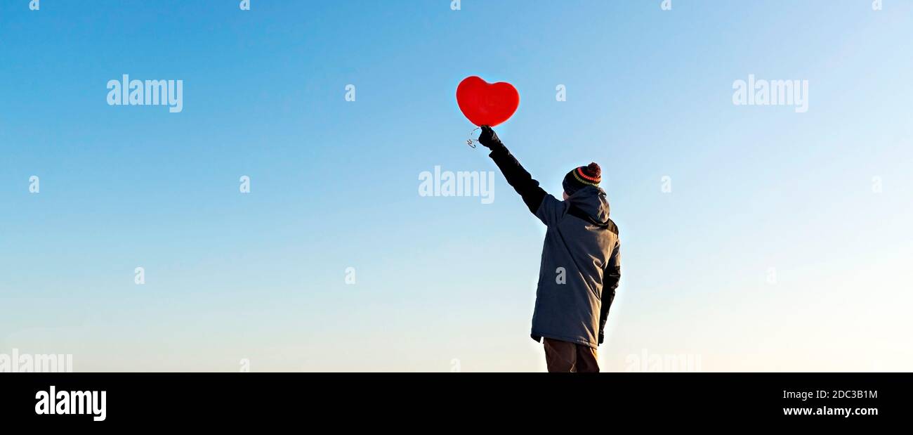 giovane uomo dalla schiena in abiti caldi che tiene un palloncino rosso a forma di cuore contro il cielo blu. San Valentino e romantico concetto, copia spazio Foto Stock