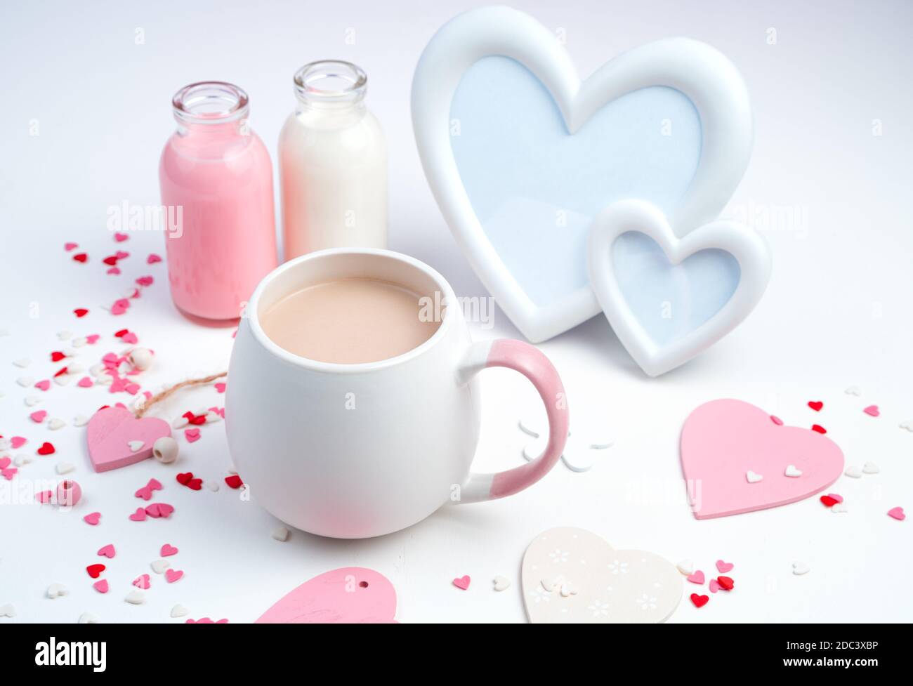 Una tazza di caffè con latte, una cornice a forma di cuore e una bottiglia di latte su fondo bianco con cuori. Foto Stock