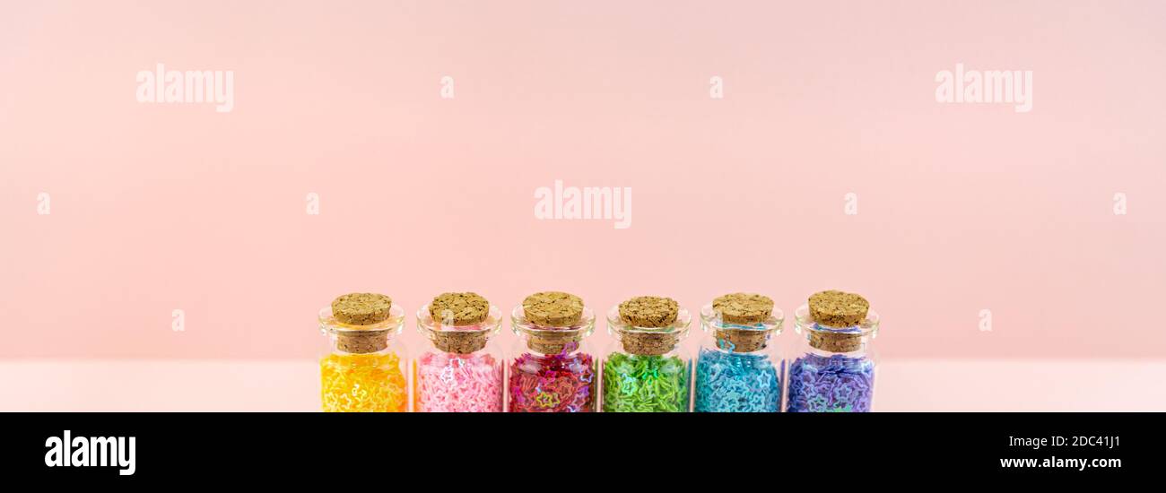 Piccole bottiglie o vasetti di vetro tappi sigillati e stelle luccicanti multicolore si trovano in fila contro uno sfondo rosa. Banner. Decorazione a paillettes Foto Stock