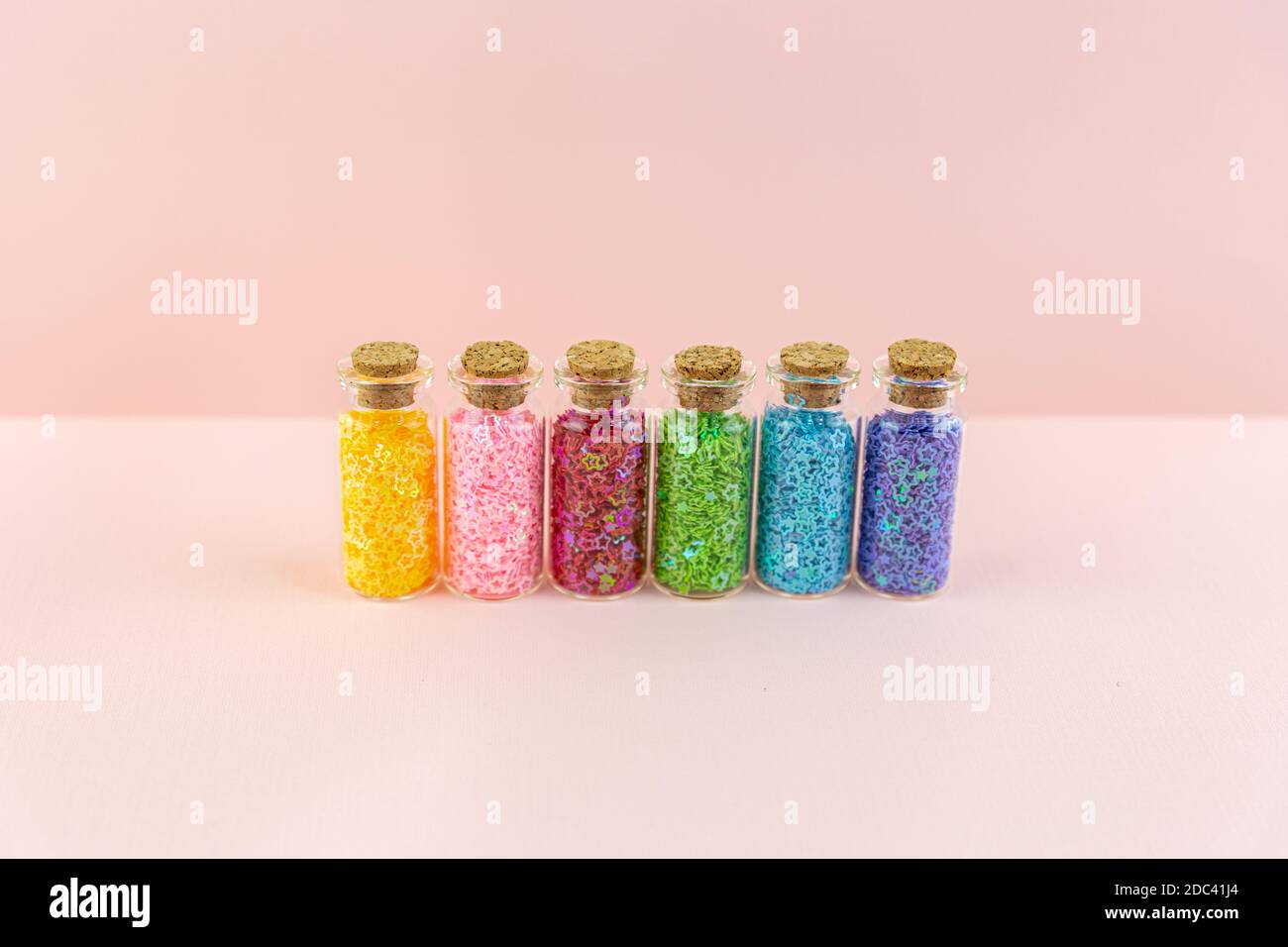 Piccole bottiglie o vasetti di vetro con tappi sigillati con stelle lucide multicolore si trovano in fila su sfondo rosa. Decorazioni in paillettes per la creatività. Foto Stock