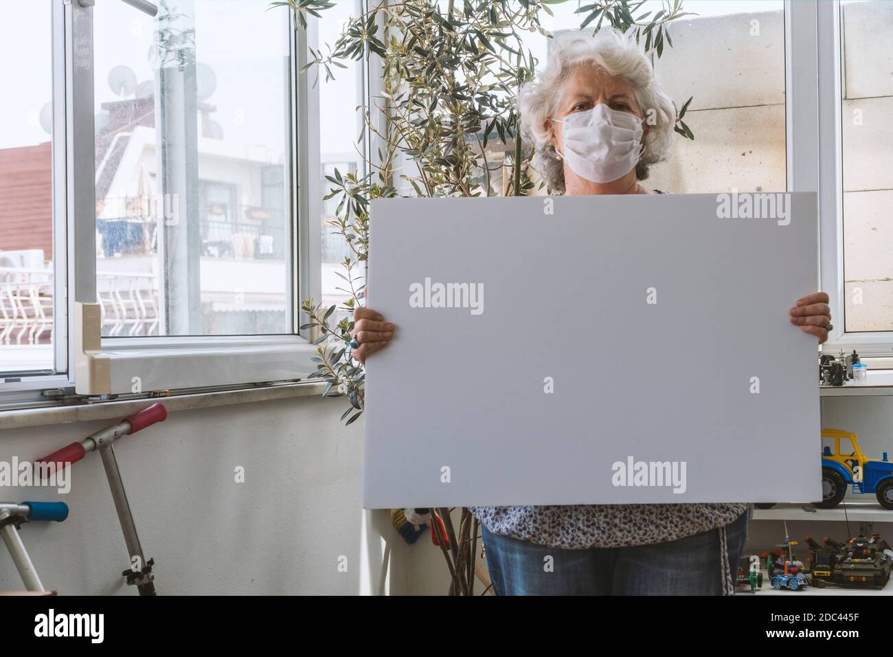 Covid-19 concetto di soggiorno a casa: Una donna anziana con i capelli grigi indossa una maschera facciale e tiene una lavagna bianca vuota senza un messaggio. È in piedi Foto Stock