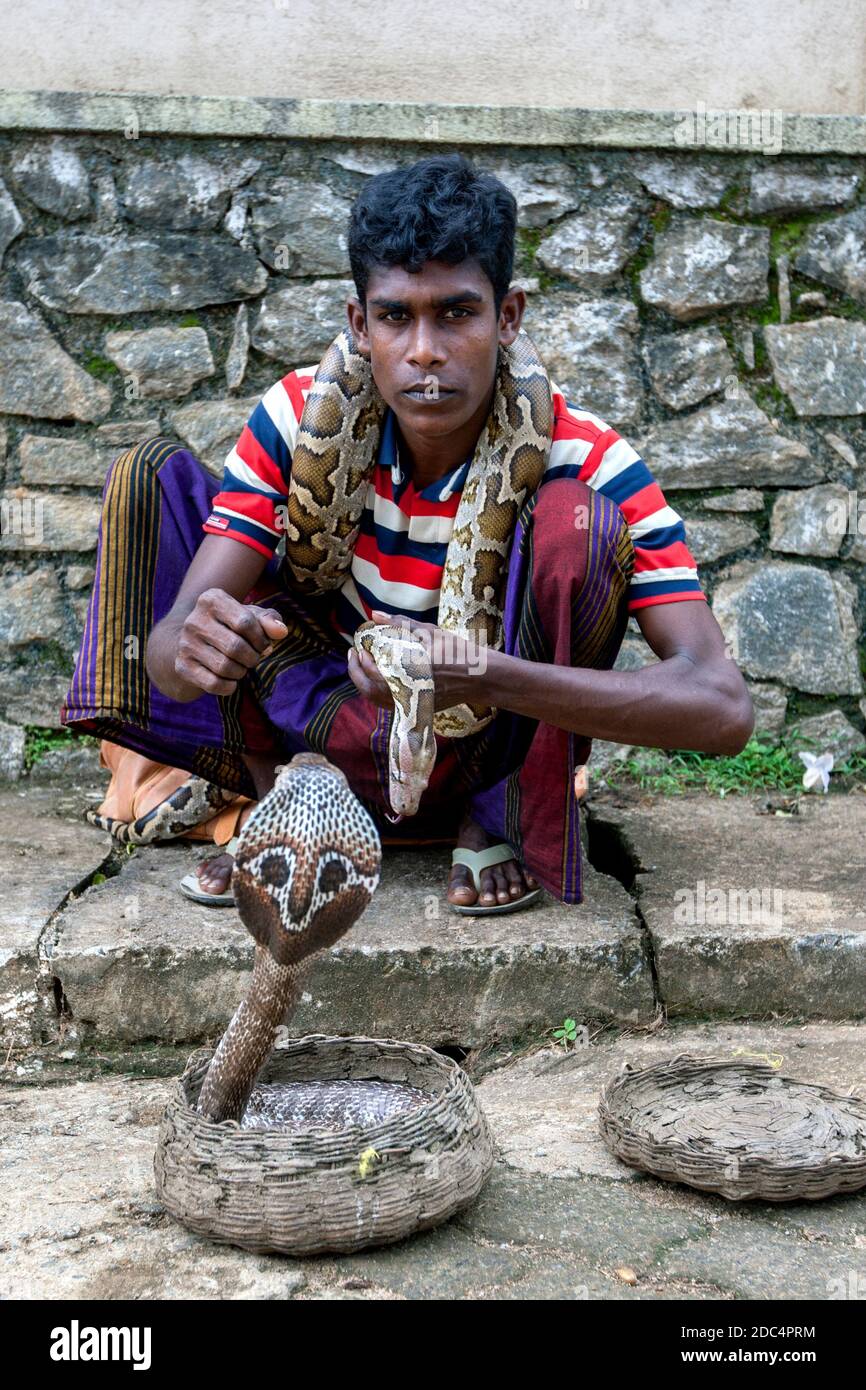 Un giovane incantatore di serpenti con un pitone avvolto intorno al collo e una cobra in un cesto di canna si trova sul sentiero a Pinnawala, nello Sri Lanka centrale. Foto Stock