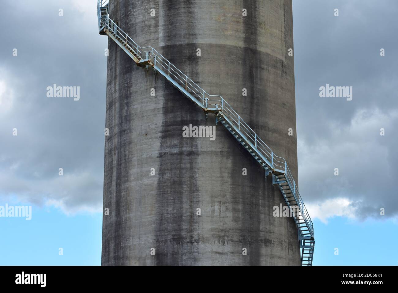 Grande silo industriale cilindrico in calcestruzzo con scala in acciaio che si avvolge sotto nuvola di tempesta scura. Foto Stock