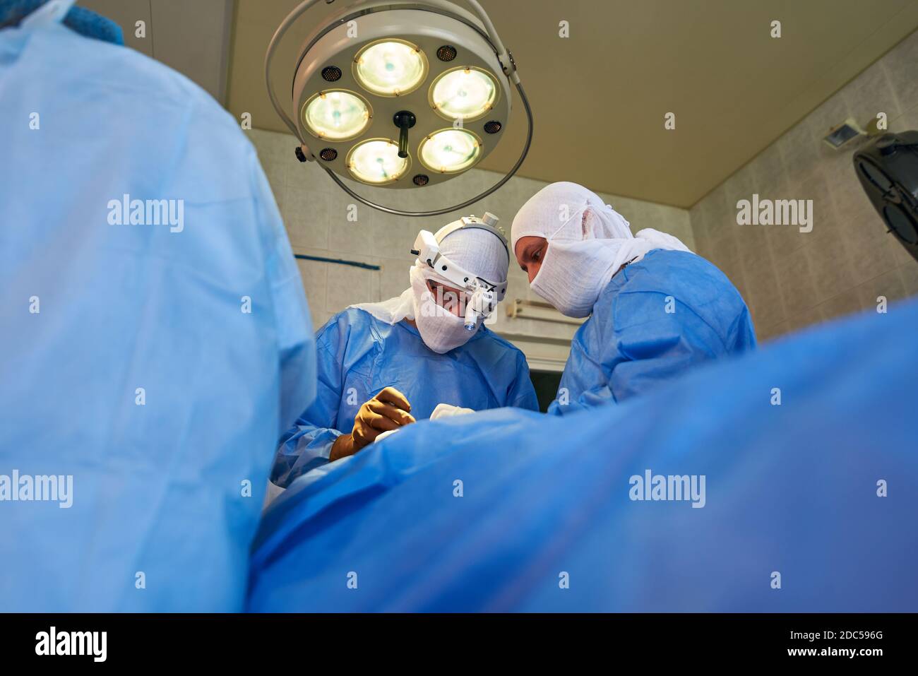 Chirurgia. Vista dal basso verso l'alto delle facce dei chirurghi sotto una lampada chirurgica rotonda. Foto Stock