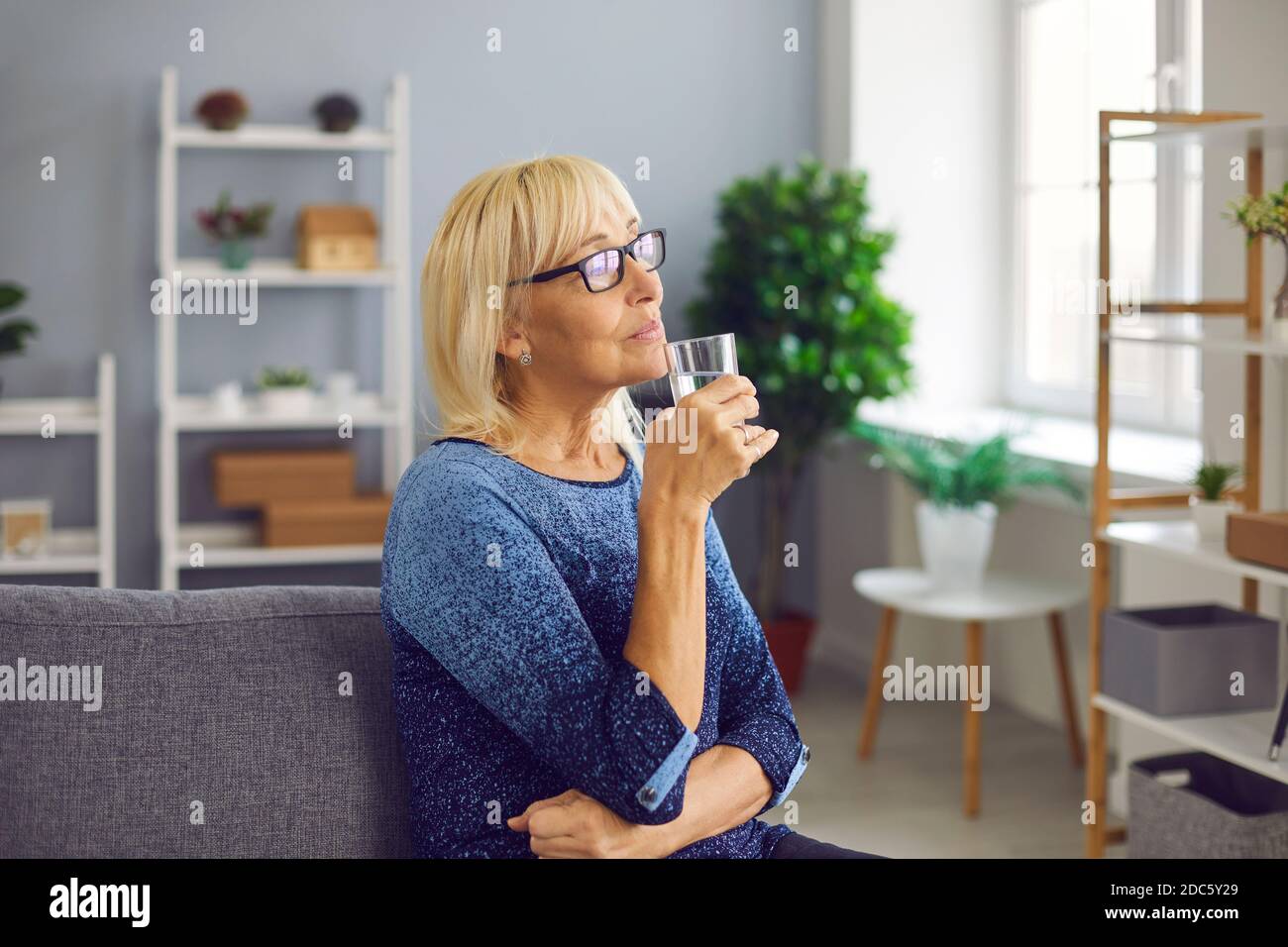 Donna anziana pensiva che beve un bicchiere di acqua pura per rimanere idratati e sani Foto Stock