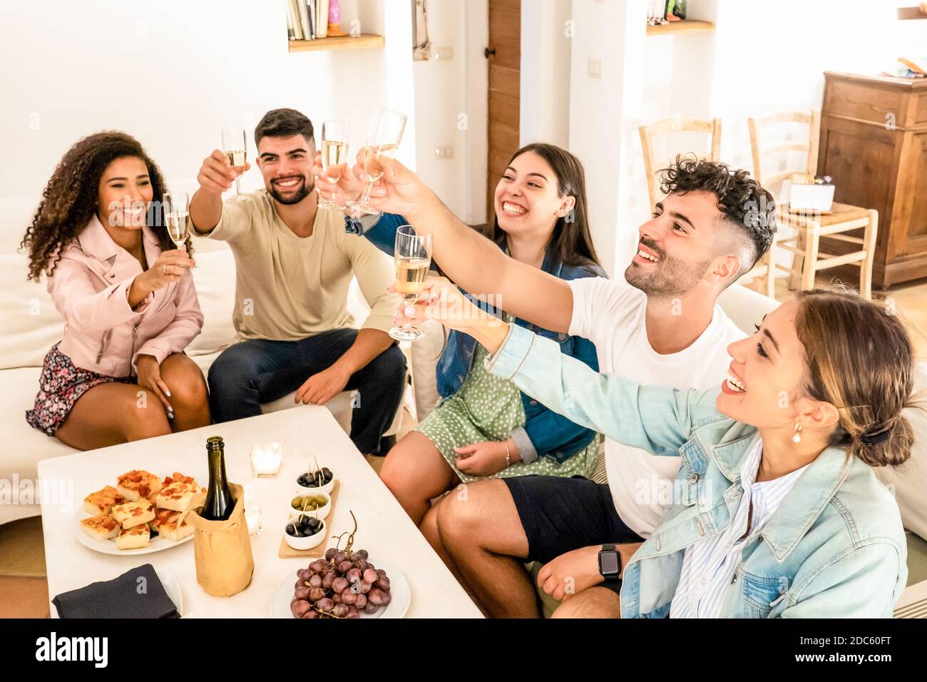 Gruppo di amici di corsa misti che tostano con champagne festeggiando a. home - giovani multirazziali seduti sul divano che si aggraffa bicchieri di vino bianco Foto Stock