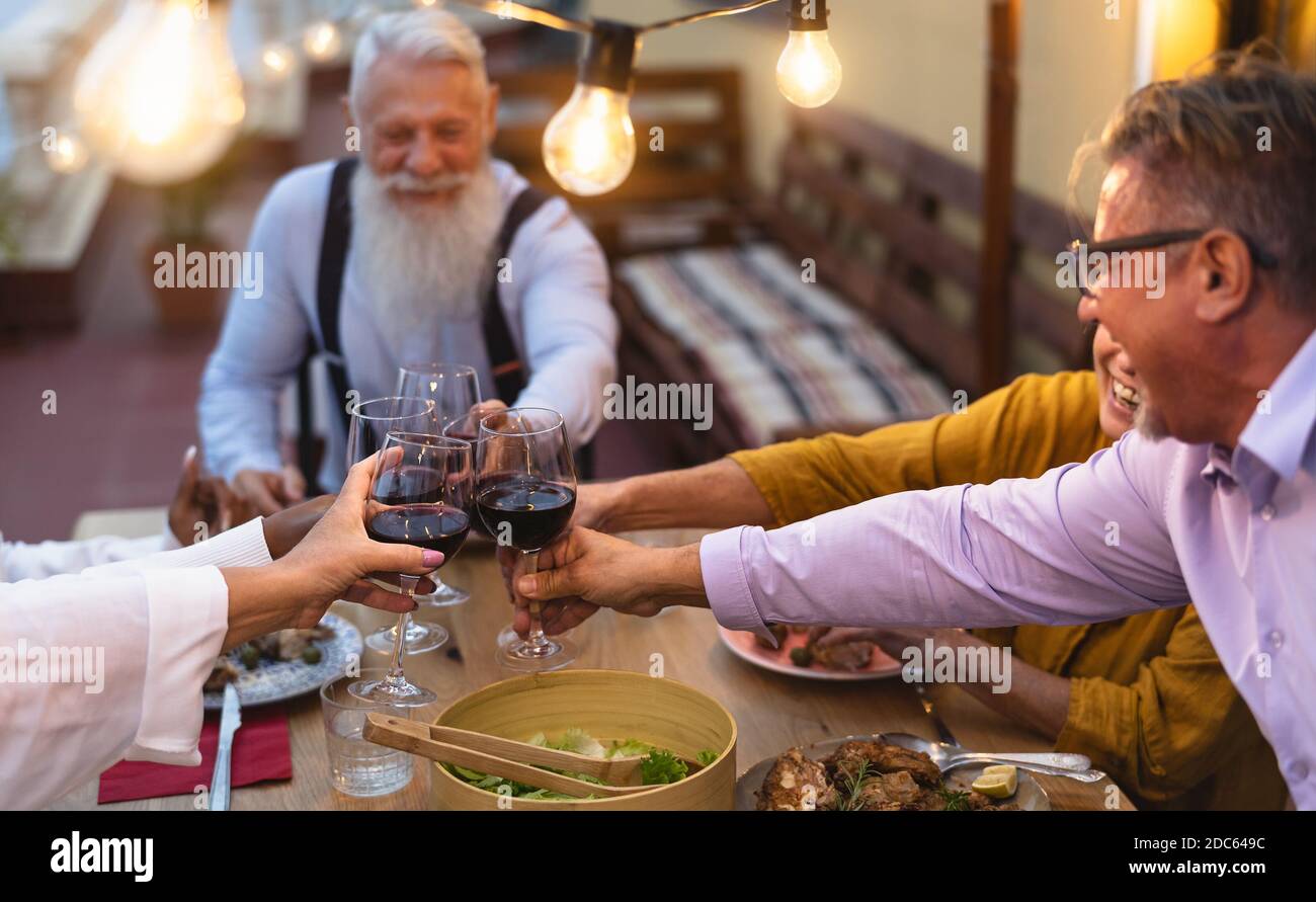 Felici amici anziani multirazziali che tostano insieme con bicchieri di vino rosso Cena nel patio della casa - anziani e cibo concetto Foto Stock