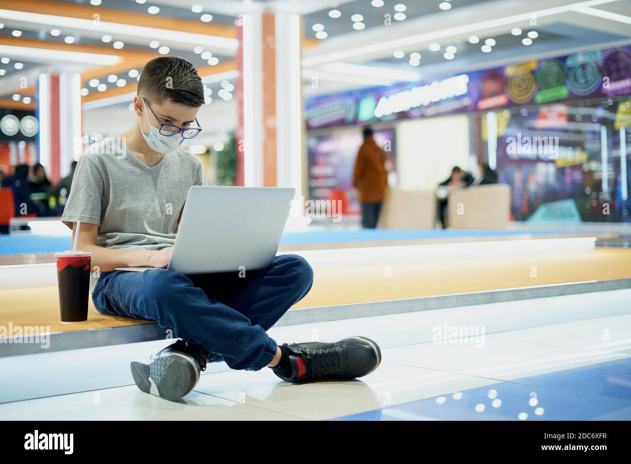 Il ragazzo teen in maschera di faccia usa il laptop nel centro commerciale Foto Stock