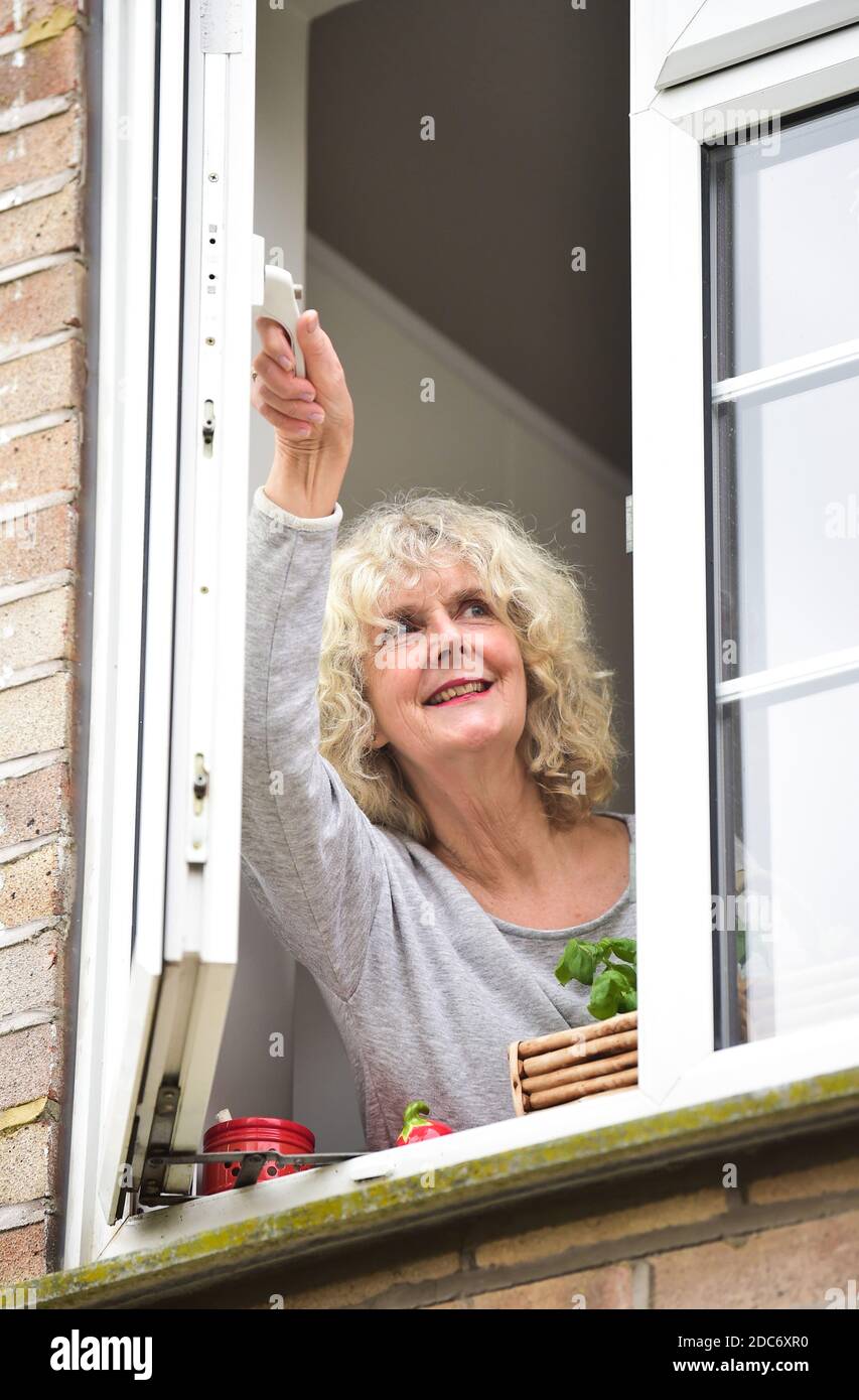 Donna a casa che apre la finestra della cucina per lasciare in fresco Air come consigliato per aiutare a combattere il virus COVID-19 del coronavirus Foto Stock