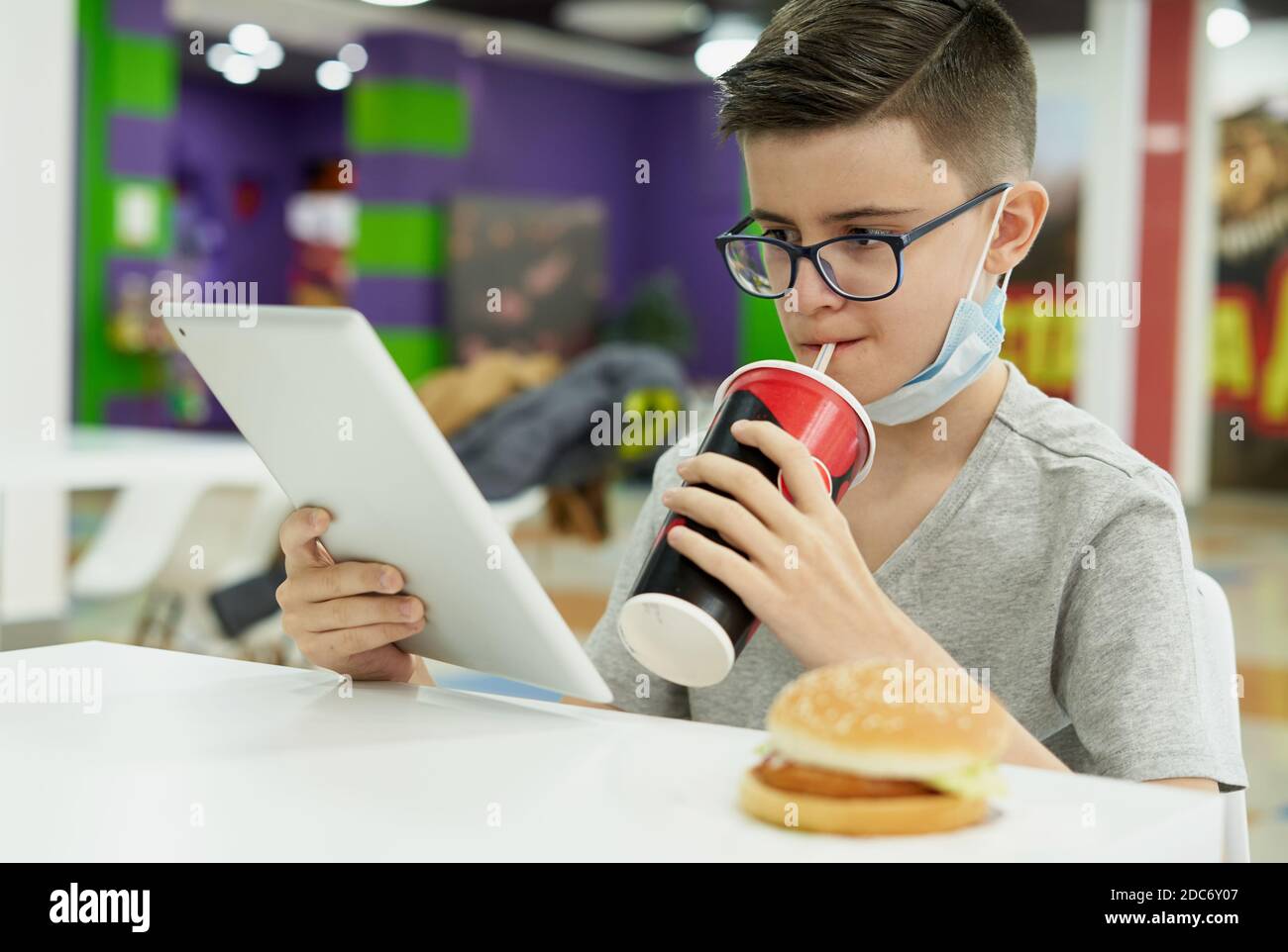 Il ragazzo adolescente toglie la sua maschera facciale e beve soda durante la riproduzione con tablet digitale Foto Stock