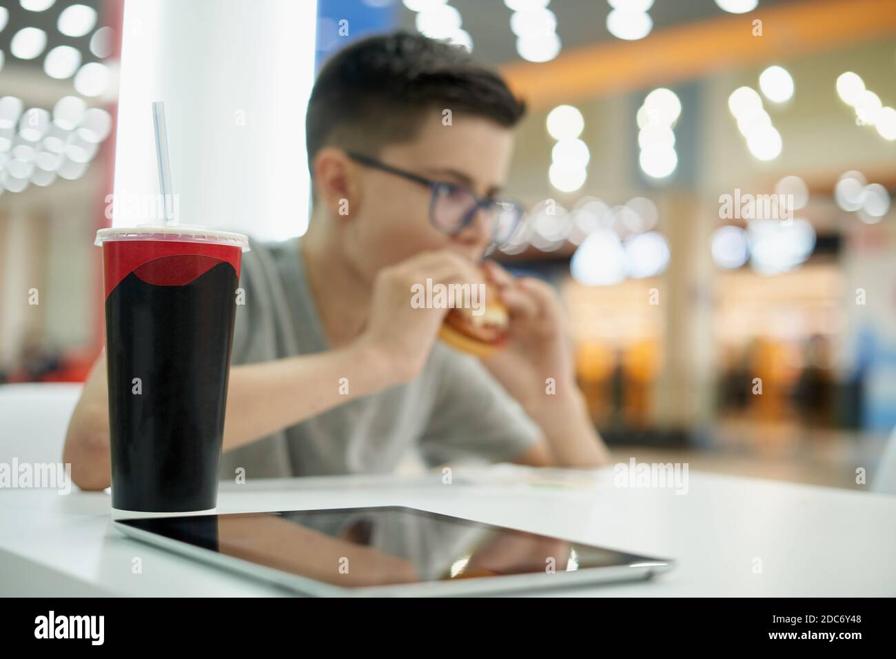 Una tazza di carta di soda e un ragazzo teenager che mangia hamburger nel ristorante fast food. Cibo malsano e adolescente Foto Stock