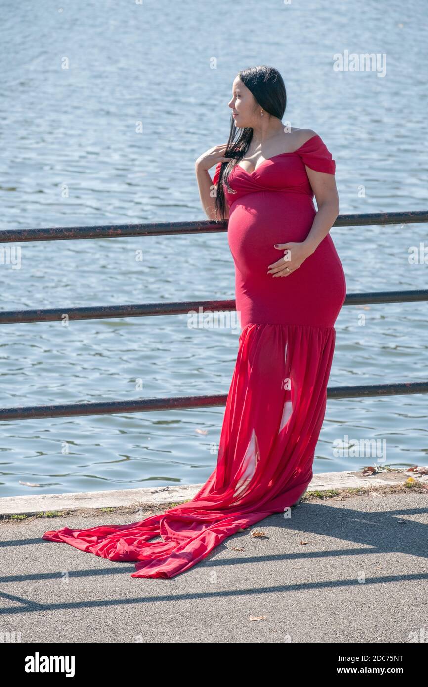 Una donna fortemente incinta in un lungo vestito scorrevole pone per le foto in un giorno d'autunno mite. Vicino a un lago in un parco a Queens, New York. Foto Stock
