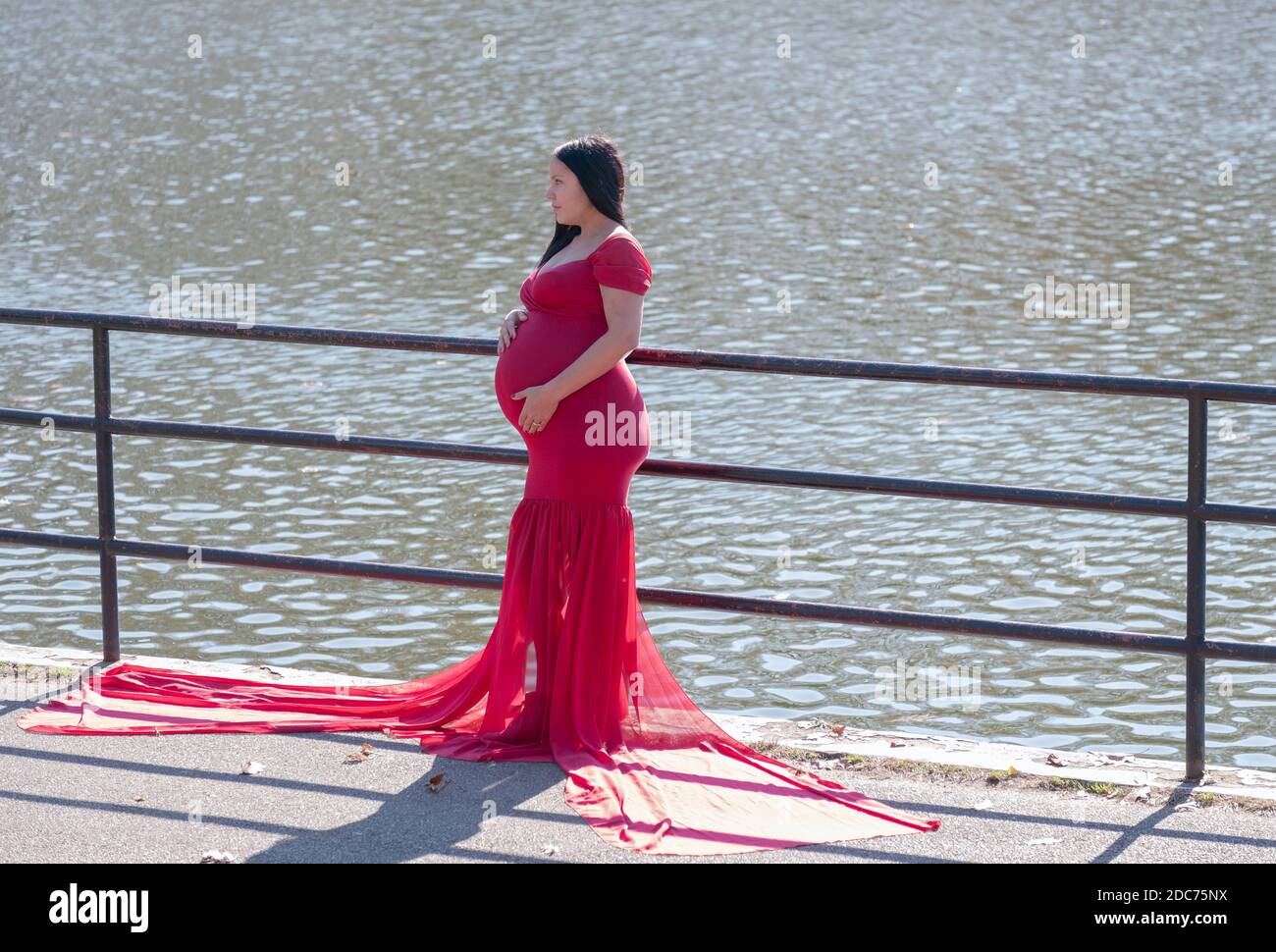 Una donna fortemente incinta in un lungo vestito scorrevole pone per le foto in un giorno d'autunno mite. Vicino a un lago in un parco a Queens, New York. Foto Stock