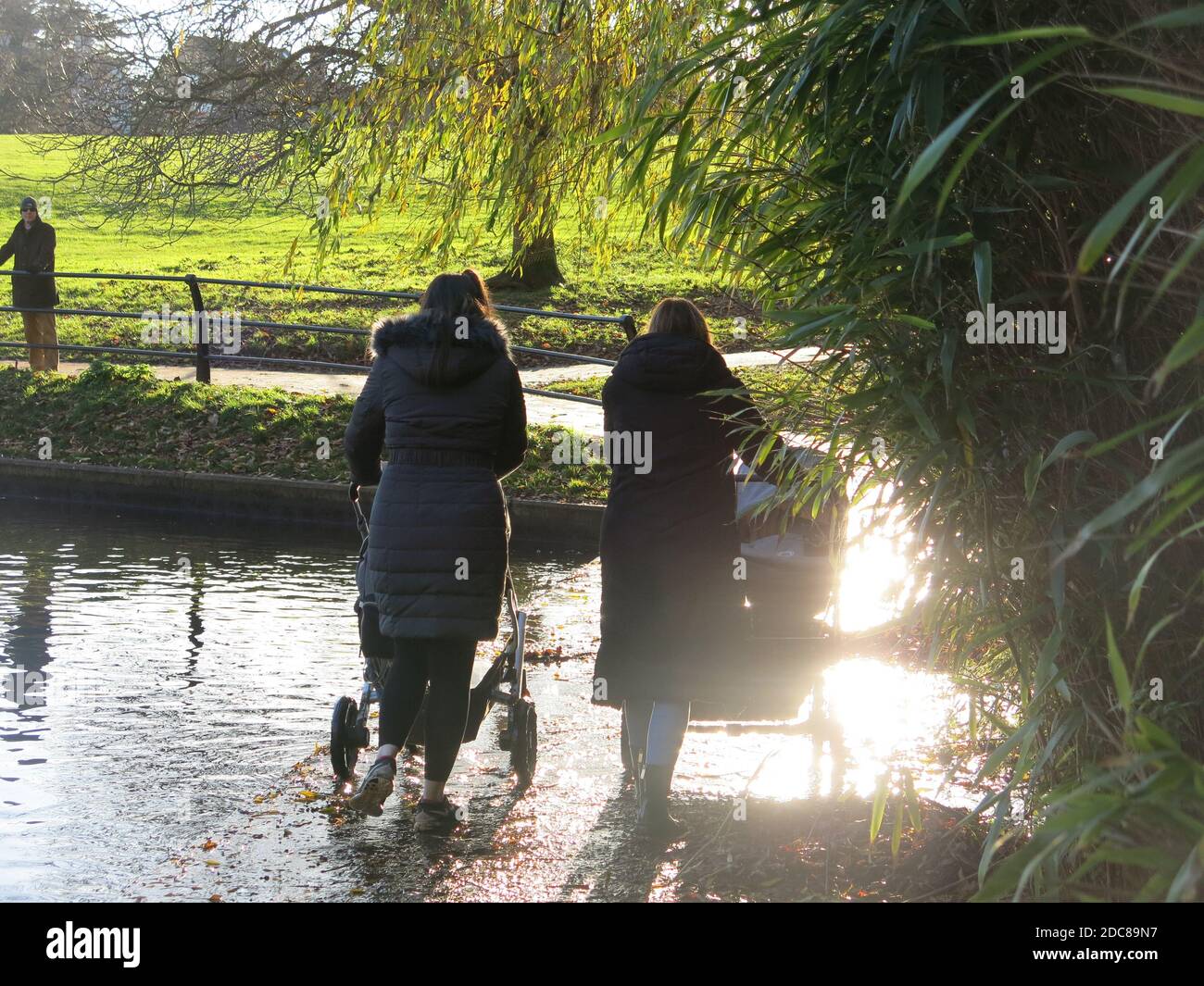 Due giovani mamme in camici invernali si sono silhouette alla luce del sole autunnale, spingendo i loro prosciutti sul sentiero allagato ai margini del lago ad Abington Park. Foto Stock