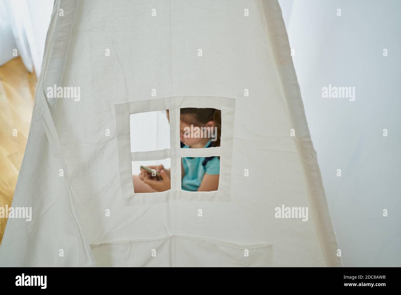 Bambina che guarda il suo smartphone all'interno di una tenda bianca in teepee all'interno della sua casa. Concetto di tecnologia Foto Stock