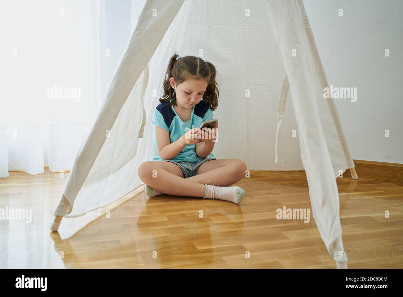 Bambina che guarda il suo smartphone all'interno di una tenda bianca in teepee all'interno della sua casa. Concetto di tecnologia Foto Stock