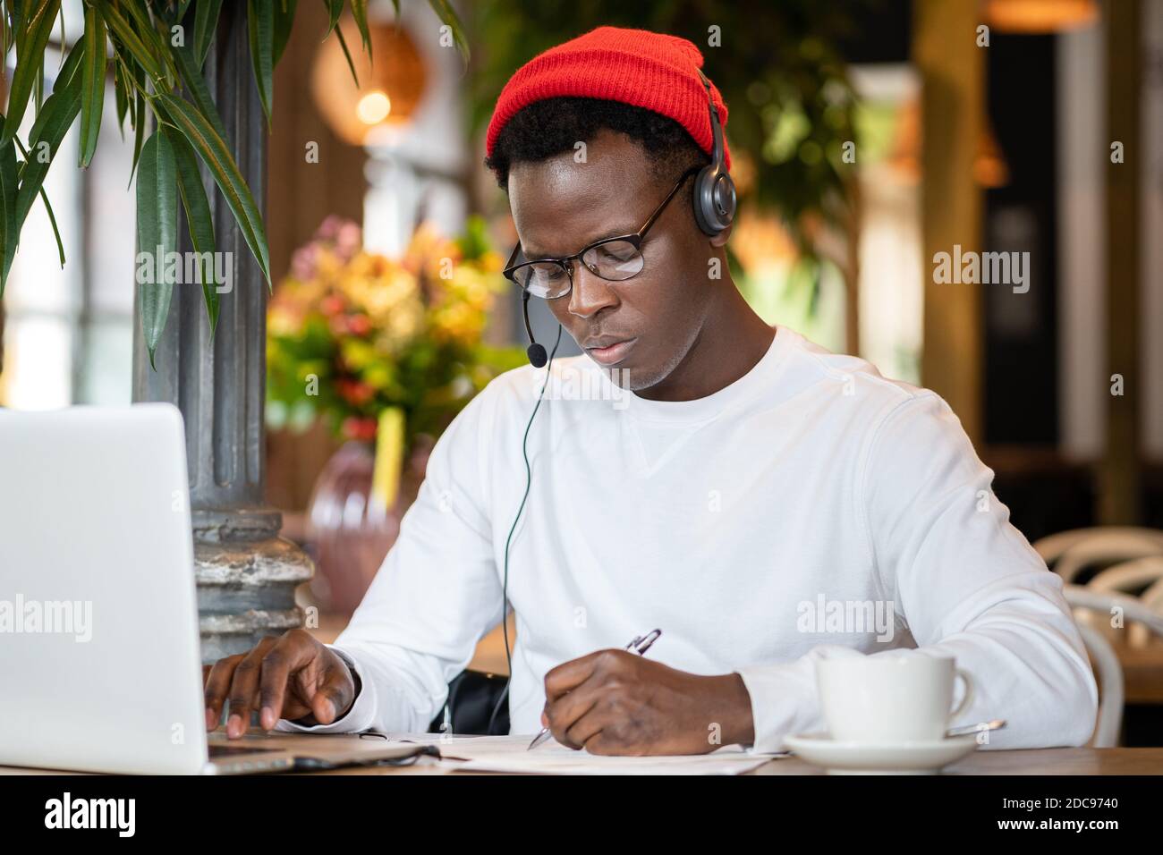 Uomo nero millenario concentrato in cuffie indossare cappello rosso, guardare webinar educativo sul computer portatile, lavorare in linea in remoto in bar, ascoltare lezione audio Foto Stock