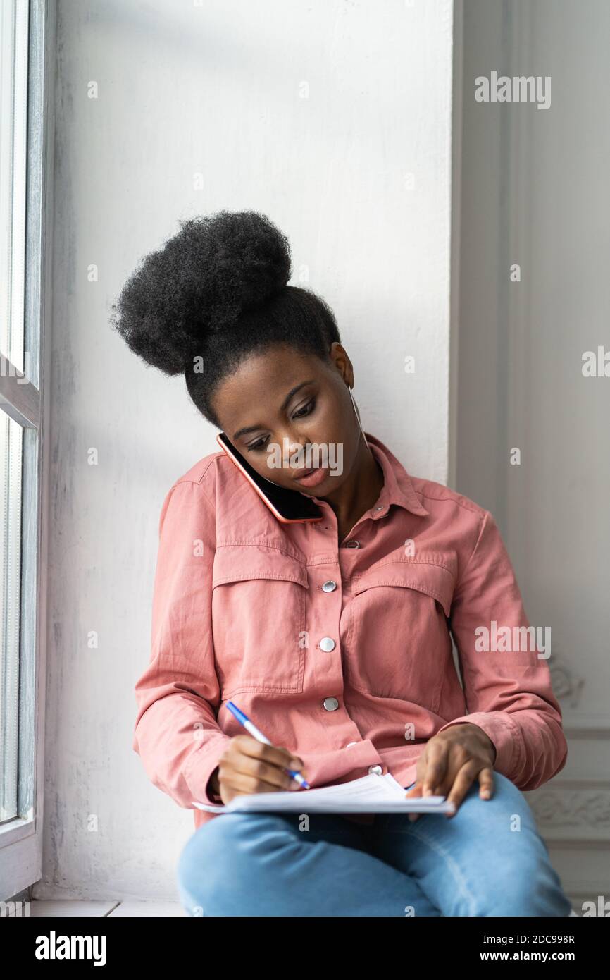 Donna biraciale afro-americana focalizzata che prende appunti, chiarisce le informazioni, parlando al telefono, seduto sul davanzale. Biracial femminile dipendente Consult Foto Stock