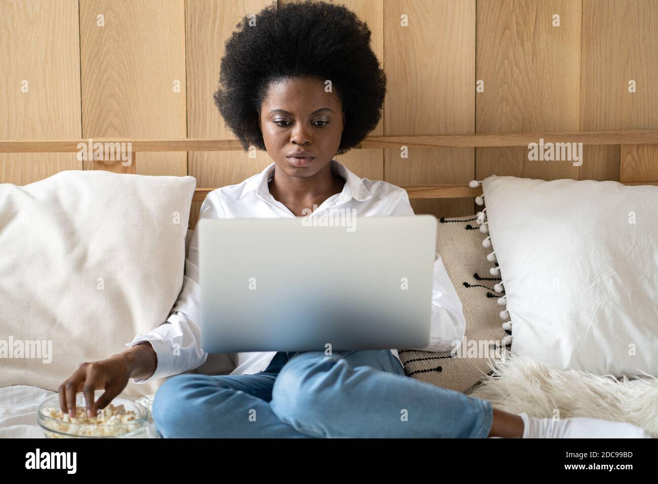 Donna afro-americana focalizzata in camicia bianca che si rilassa dopo il lavoro, mangiando popcorn mentre guardi film horror o thriller sul laptop, sdraiato a letto. Lazy r Foto Stock