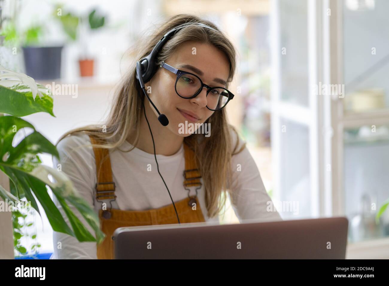 La donna millennial del giardiniere indossa le cuffie usando il laptop, comunica con i clienti, guarda il webinar o videoconferenza in streaming, lavora a distanza da casa Foto Stock