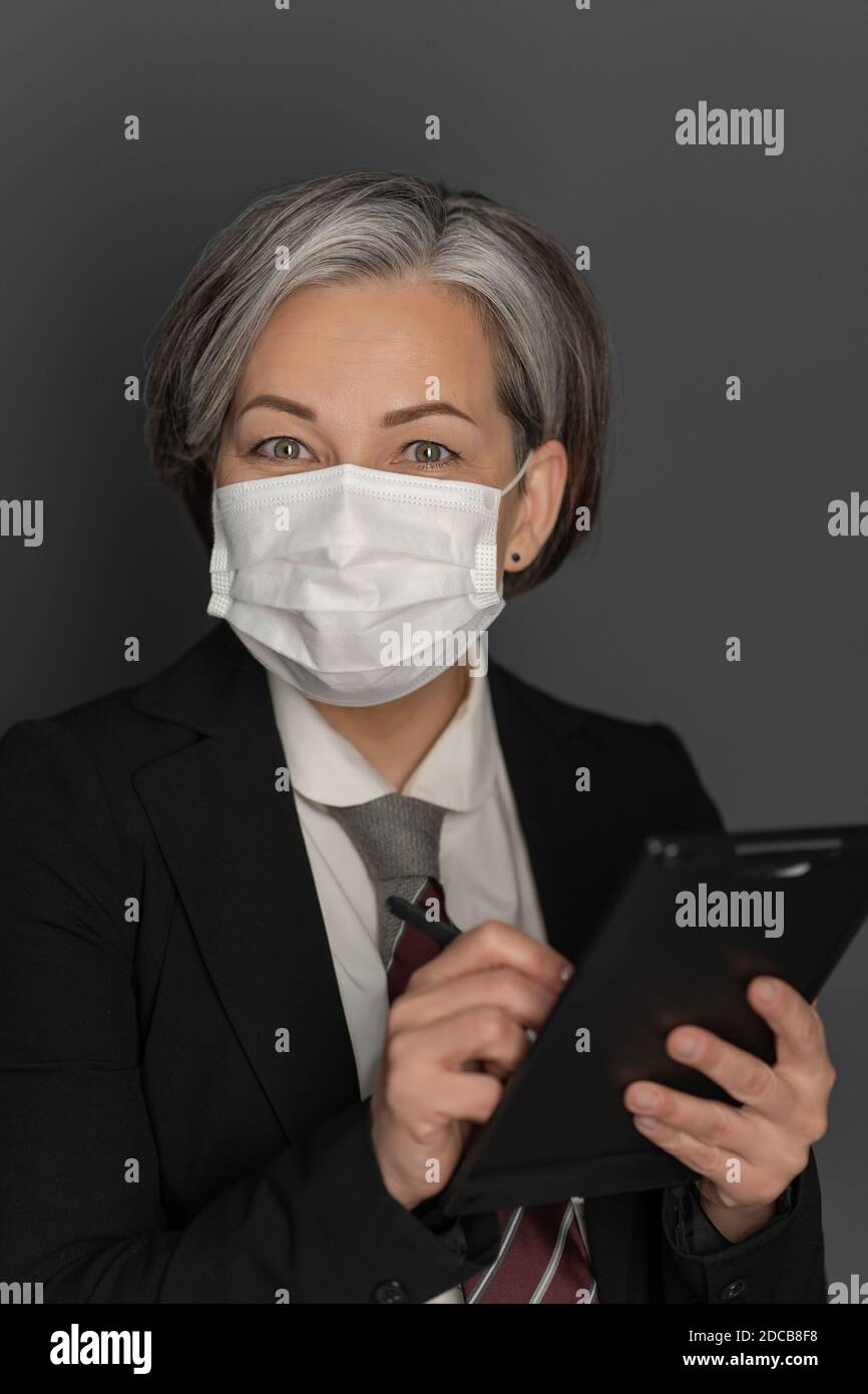 Donna d'affari con capelli caucasici grigi in maschera protettiva lavoro utilizzando il gadget tablet digitale guardando la fotocamera isolata su sfondo grigio. Scatto in studio Foto Stock