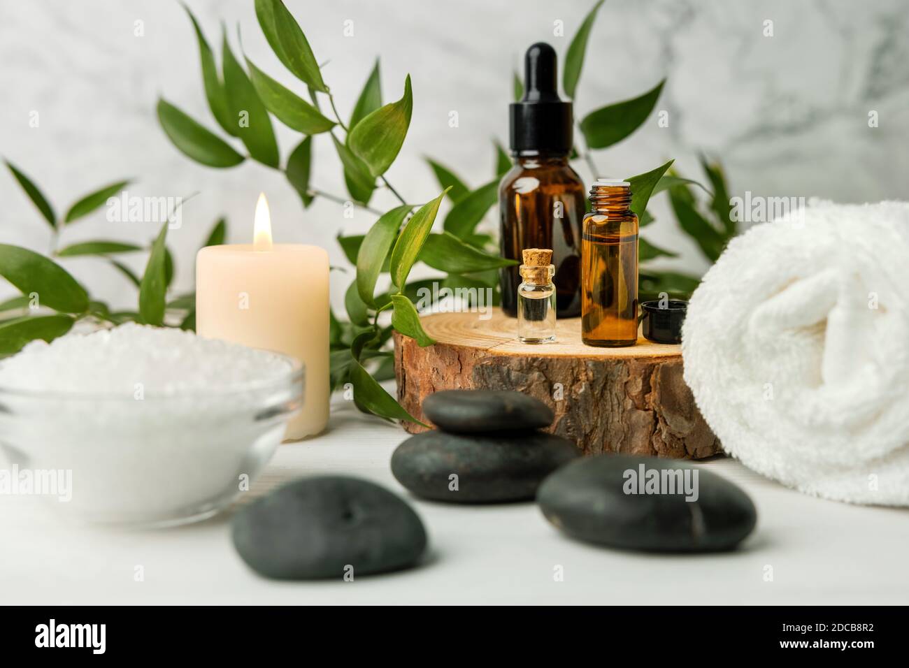 articoli per trattamenti di bellezza per trattamenti termali su tavola di legno bianco con piante verdi, pietre da massaggio, oli essenziali e sale marino con candela bruciante Foto Stock