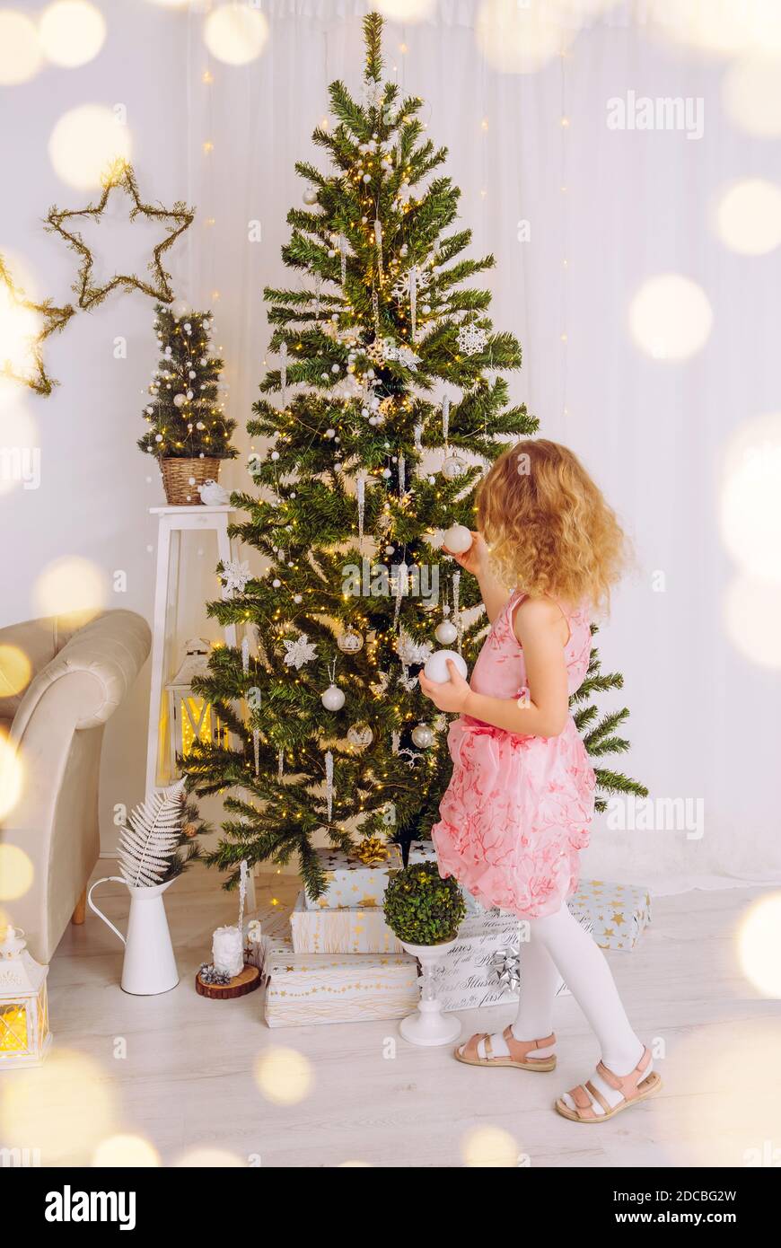 Irriconoscibile bambina di 6 anni in abito da festa rosa, appeso ornamenti di Natale bauble sull'albero di Natale. Soggiorno bianco minimalista. Foto Stock