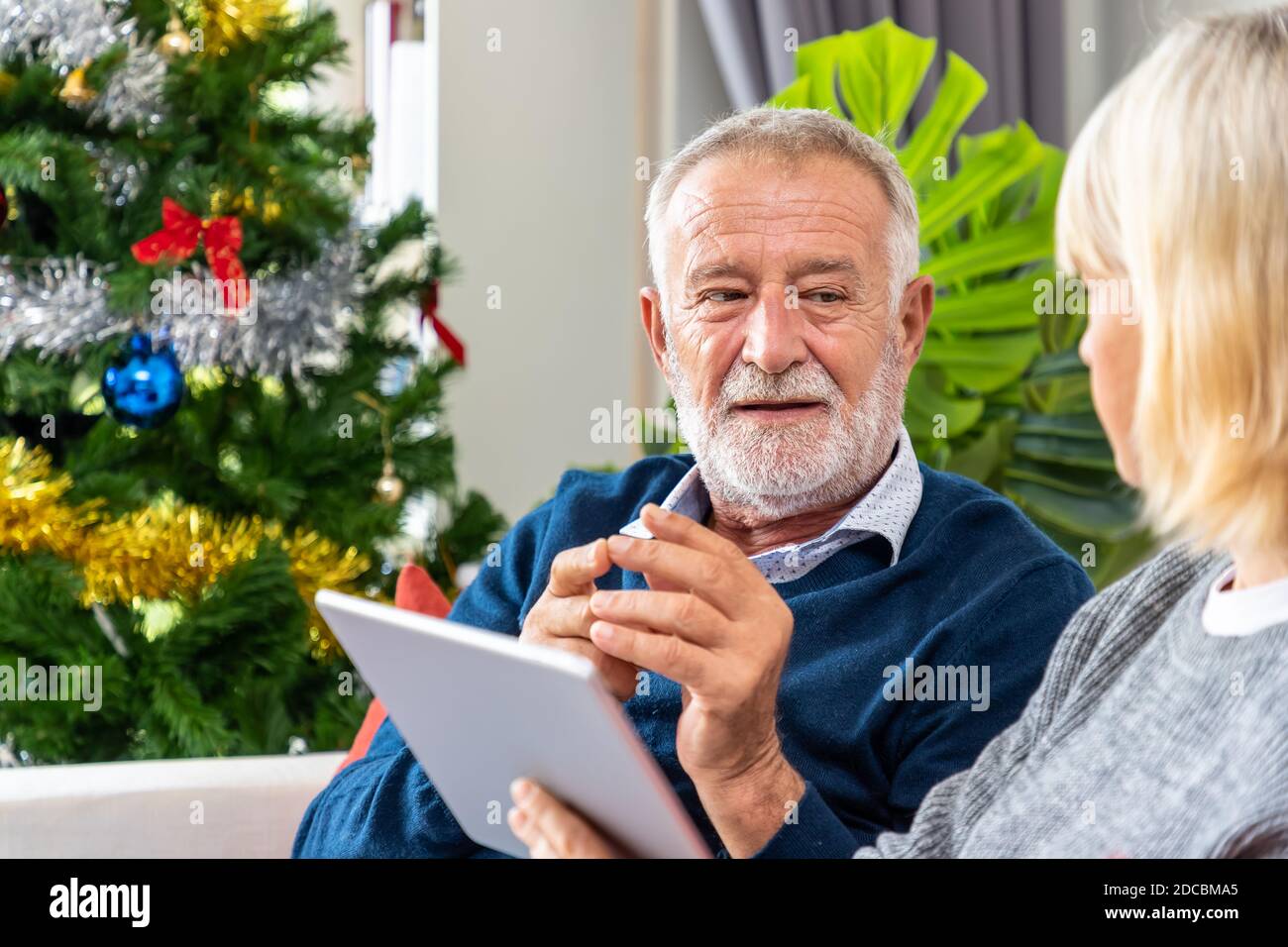 Coppia anziana che usa il tablet per videochiamare per salutare la famiglia per il festival di Natale, seduto sul divano con decorazione e albero Foto Stock
