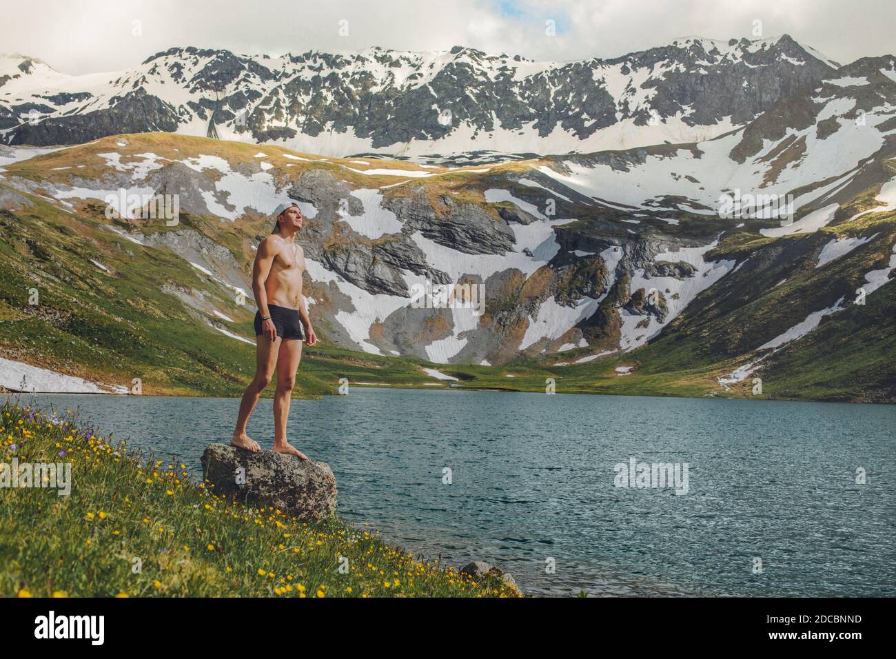 Giovane turista maschile in un costume da bagno che si erge sulla riva di un lago di montagna puro e gode della natura sullo sfondo di montagne innevate. Foto Stock