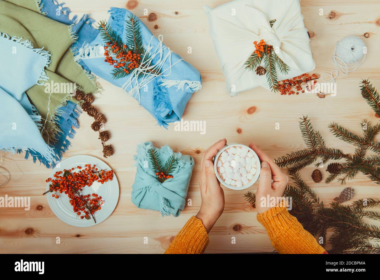 Le mani femminili tengono una tazza di cococa con marshmallow e appena imballate scatole regalo avvolte in tessuto e decorate con ramo di abete rosso, bacche giacenti sopra Foto Stock