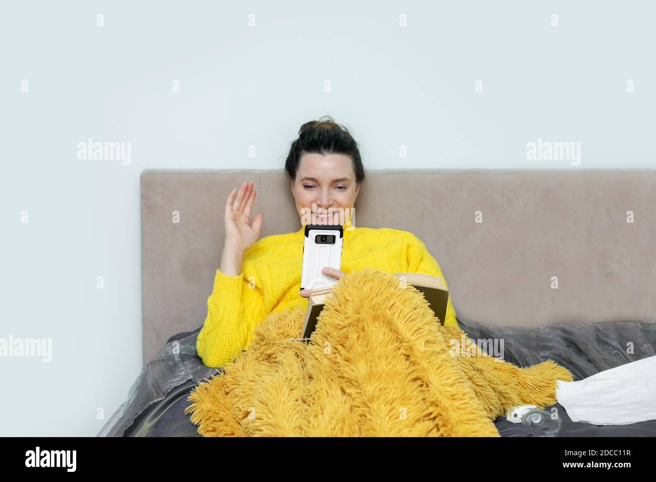Ammalato cute donna sul letto in maglione, sotto coperta in camera da letto felicemente comunica in videochiamata via smartphone. Autoisolamento durante la pandemia. Concetto Foto Stock