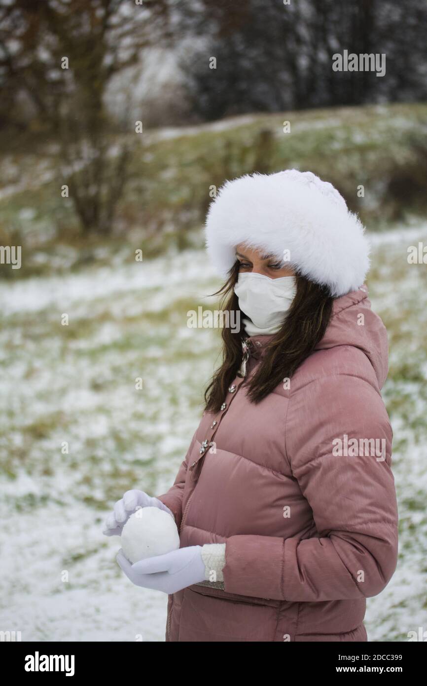 Una ragazza in una maschera medica per una passeggiata. Tiene una palla di neve nelle sue palme. Il terreno è coperto dalla prima neve Foto Stock