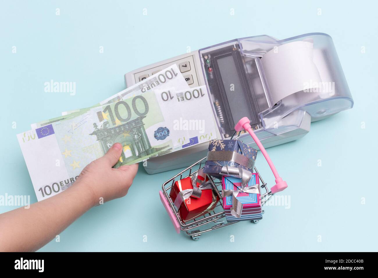 Primo piano di una mano di un bambino che tiene una copia di banconote da 100 euro sopra il registratore di cassa per l'acquisto di scatole di regali in un carrello su sfondo blu, t Foto Stock