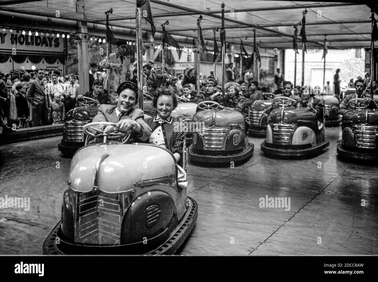 Una coppia adolescente sorride mentre guidano la loro macchina del dodgem davanti alle vetture seguenti, ad una fiera negli anni cinquanta Foto Stock