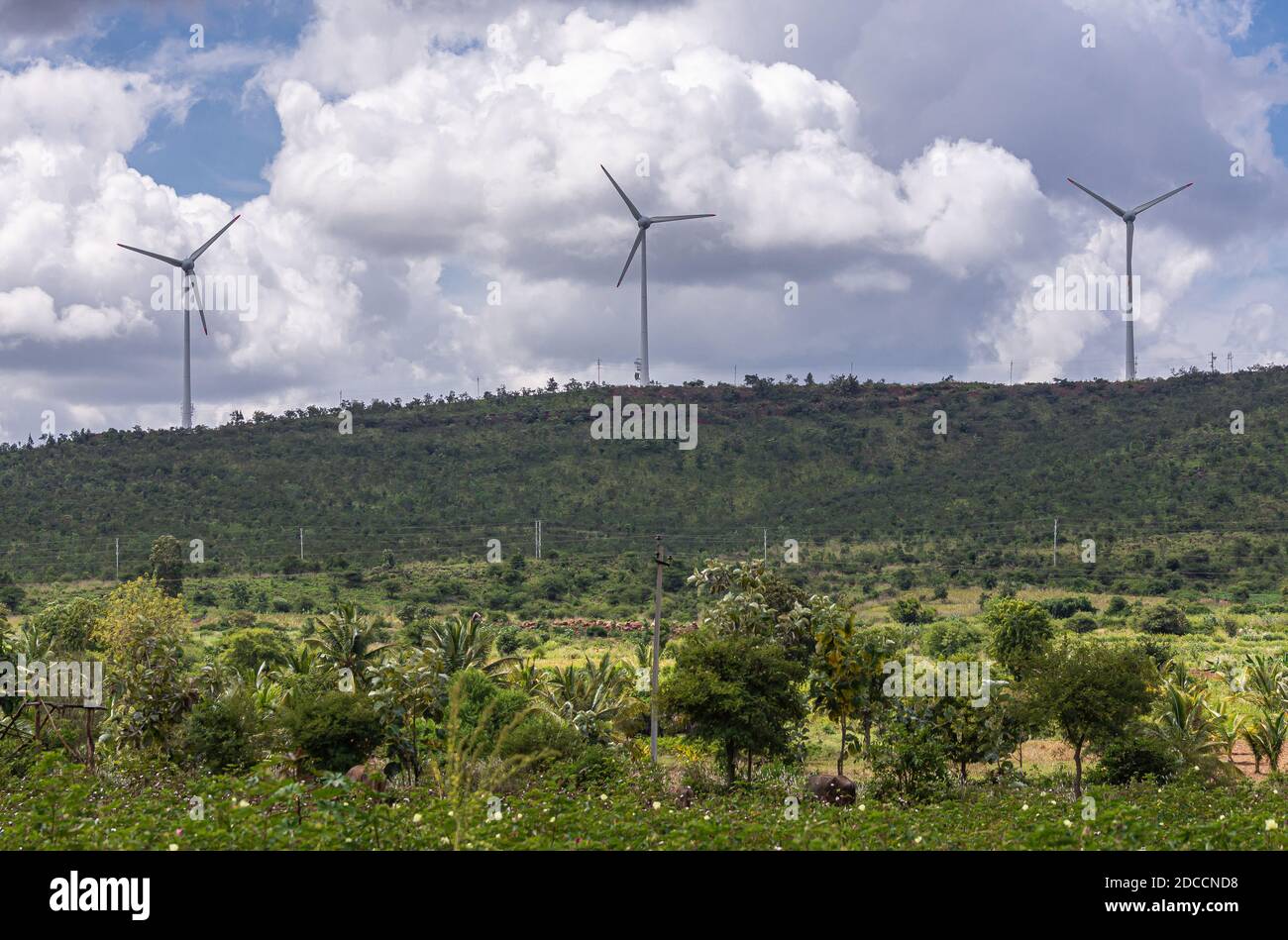 Arasinagundi, Karnataka, India - 3 novembre 2013: Paesaggio rurale verde agricolo con collina e 3 moderni mulini a vento sotto il paesaggio blu nuvoloso. Foto Stock