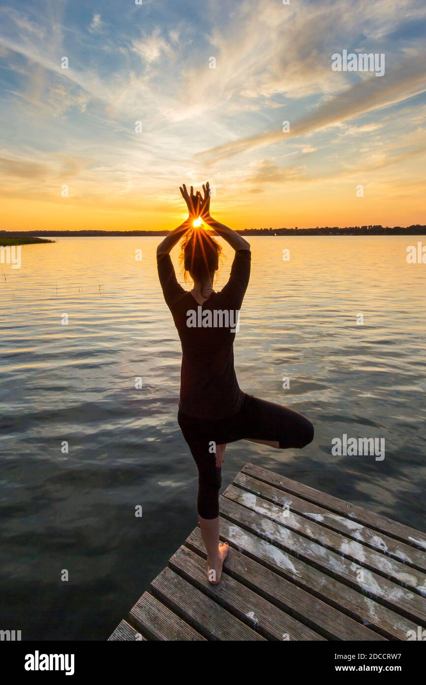 Donna che pratica la postura in piedi Vrikshasana / posa albero, una zampe di equilibrio asana della hatha medievale yoga sul molo al lago al tramonto Foto Stock