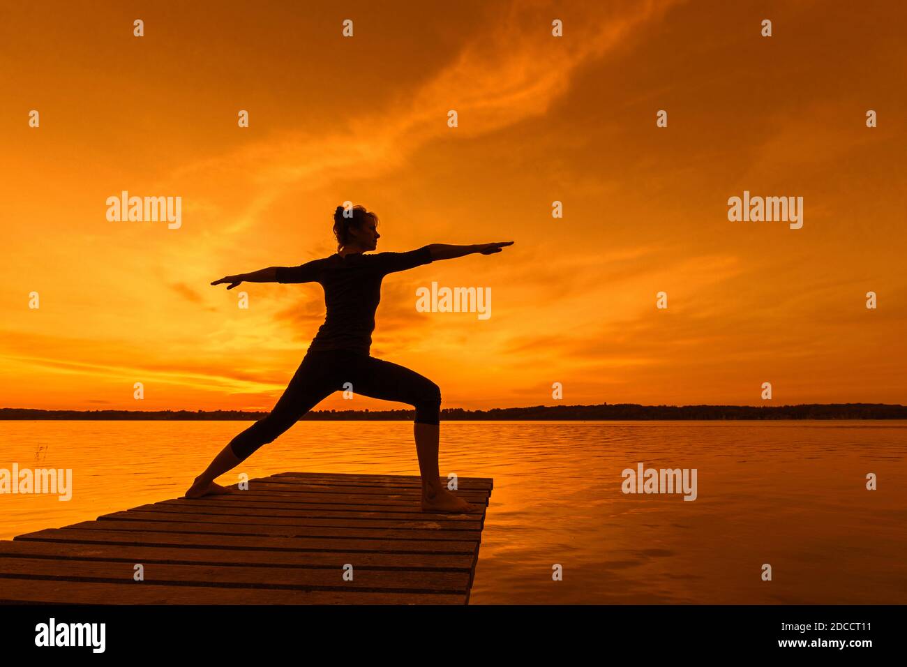Donna che pratica yoga postura Virabhadrasana II / Guerriero posa, affondo in piedi asana sul molo al lago al tramonto Foto Stock