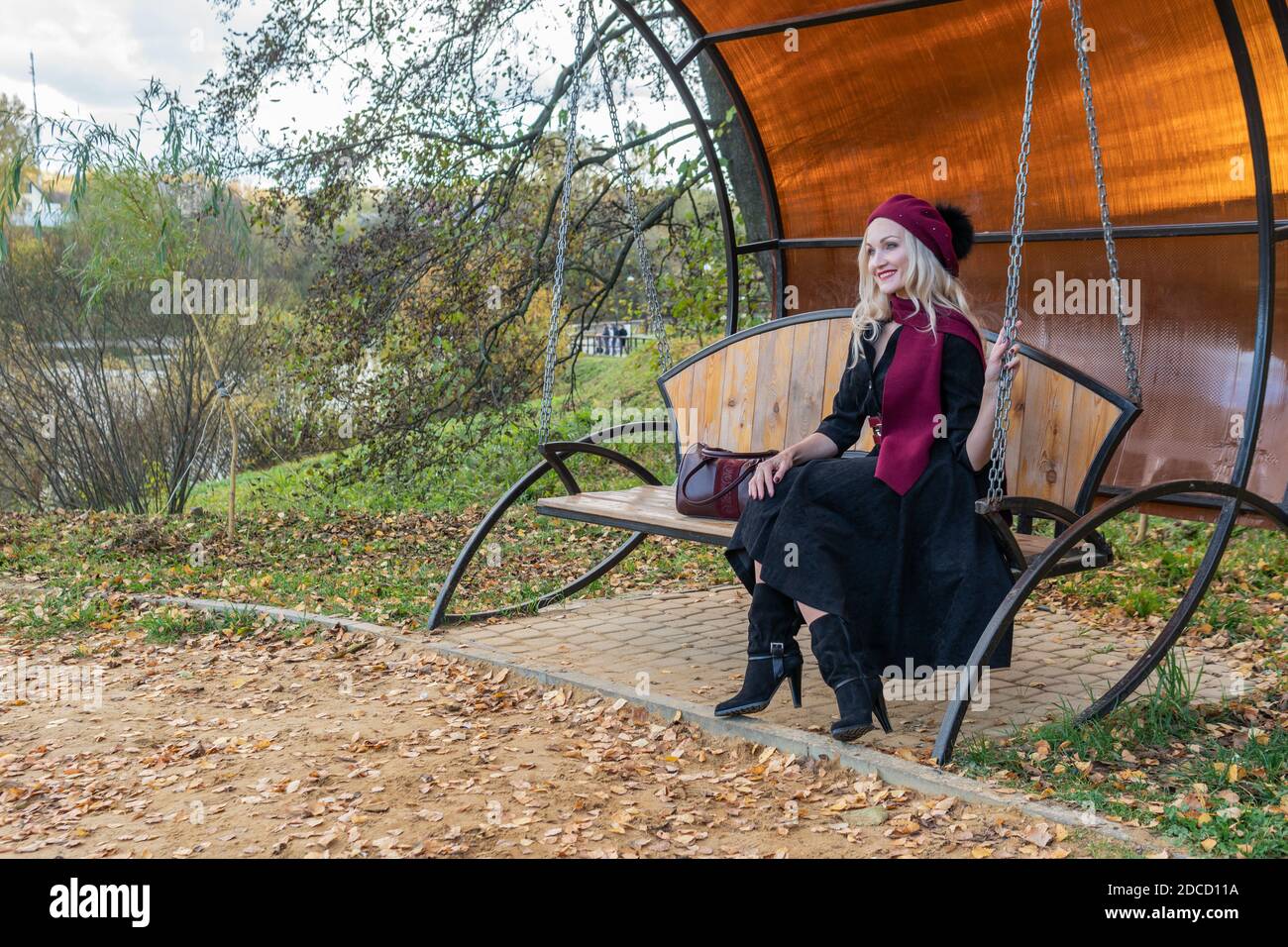 Una signora allegra si siede su un'oscillazione del giardino, forgiata in un cappotto di borgogna e biret, con gli occhi bei, nella caduta contro lo sfondo di un laghetto lì Foto Stock