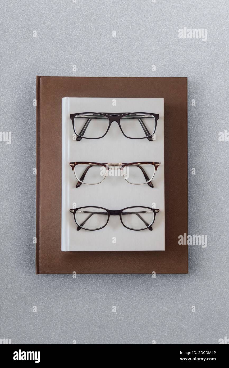 Tre diversi tipi di occhiali sulla copertina rigida di un libro grigio su una grande copia in pelle marrone su un elegante tavolo grigio testurizzato. Foto Stock