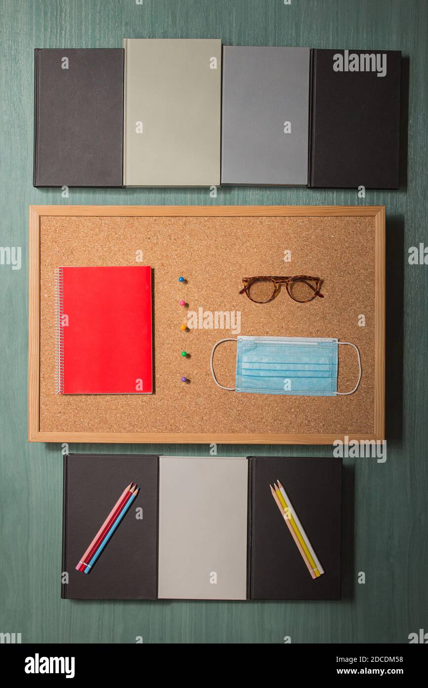 Corkboard con occhiali e un taccuino rosso separato da spille colorate e circondato da diversi libri su un tavolo di legno verde. Ritorno a scuola. Foto Stock