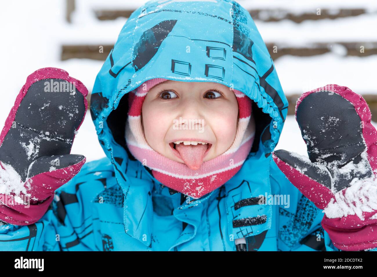 Primo piano ritratto di allegro bambino caucasico in abiti invernali. Una ragazza di 6-7 anni in una tuta invernale e mittens mostra la lingua e le palme. Wint Foto Stock