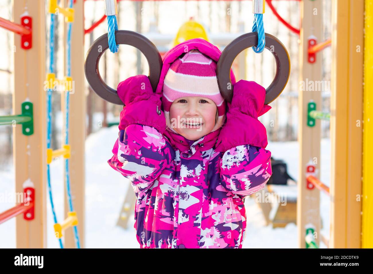 Un bambino caucasico allegro con guance rude in abiti invernali si trova sul parco giochi, guarda la macchina fotografica. Ritratto a mezza lunghezza di una bambina in inverno Foto Stock