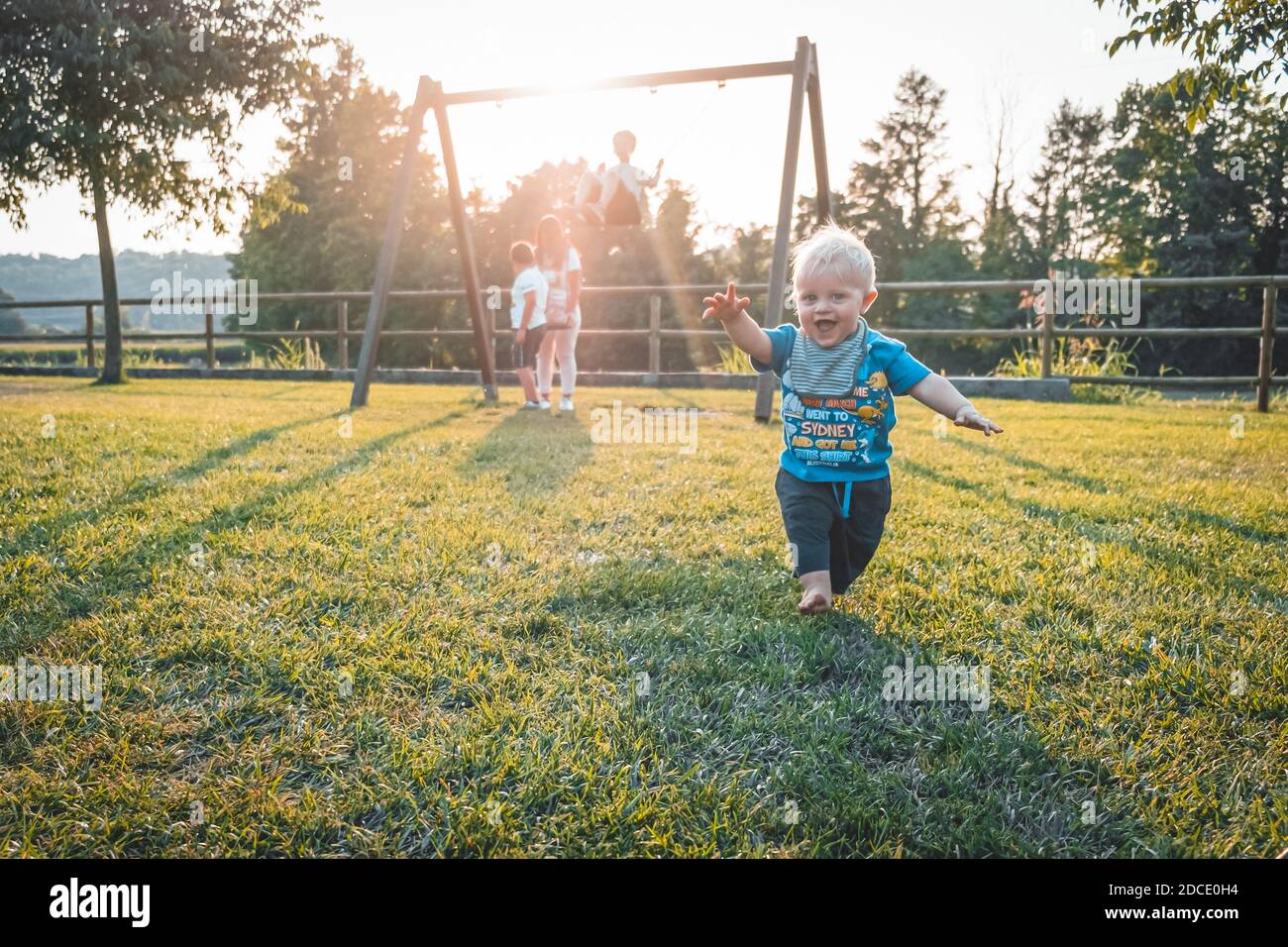 Un ragazzo di un anno che corre a piedi nudi nel parco giochi su Un bel giorno di primavera - carino ragazzino che lo prende Primi passi al tramonto - imparare a camminare conce Foto Stock